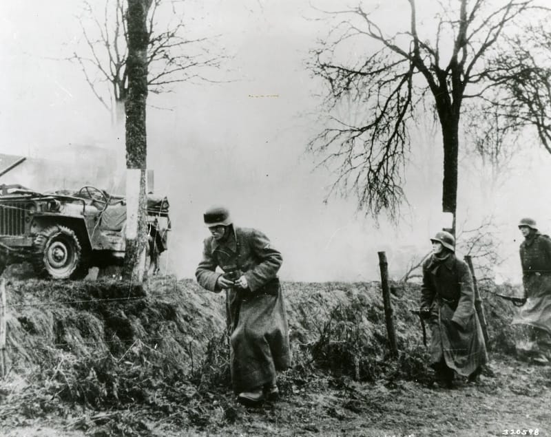 German infantry advances past an abandoned American Jeep during the Battle of the Bulge.