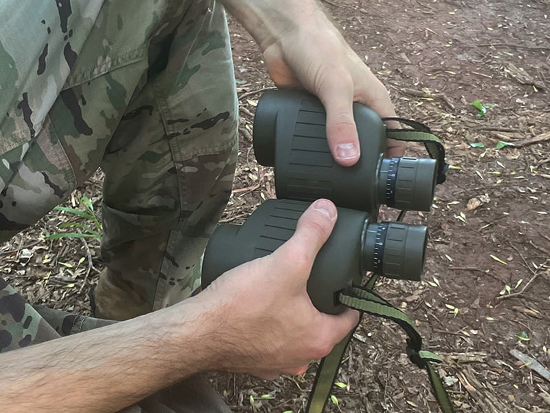 man holding binoculars with both hands, squatted to the ground